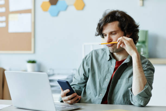 Man sitting at a desk with a laptop and smartphone, holding a pencil to his mouth and looking at his phone.