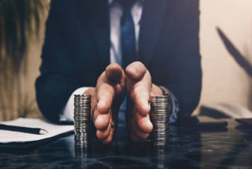 Picture2 Businessperson in a suit cupping two stacks of coins on a desk, suggesting saving or protecting money.