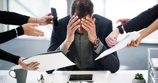 Stressed man at a desk with his head in his hands as coworkers push papers and phones toward him in a tense meeting.