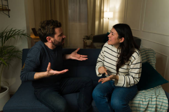 Man and woman sit on a couch in a cozy living room, mid-conversation with animated hand gestures.