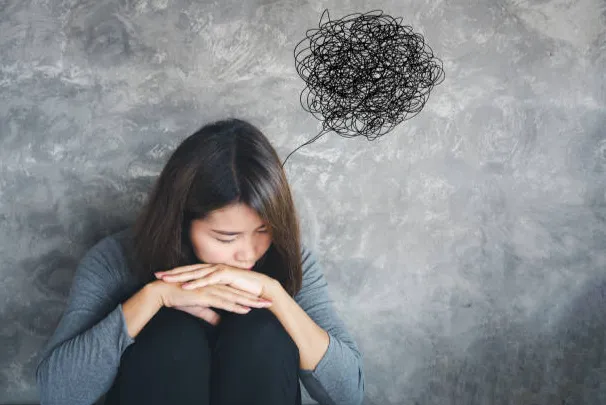 Woman sitting with knees drawn up, head bowed on folded arms, gray wall behind; a tangled scribble above her head indicating worry or confusion.